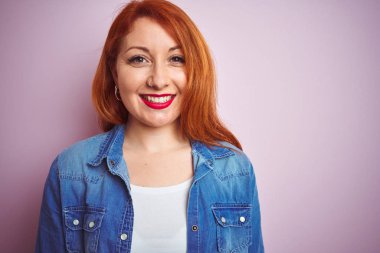 Youg güzel kızıl saçlı kadın yüzünde mutlu ve serin bir gülümseme ile izole pembe arka plan üzerinde duran denim gömlek giyiyor. Şanslı kişi.