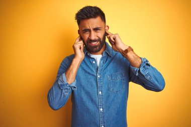 Denim gömlek giyen genç hintli adam yüksek sesle müzik sesi için rahatsız ifade ile parmakları ile kulakları kapsayan izole sarı arka plan üzerinde duran. Sağır kavramı.