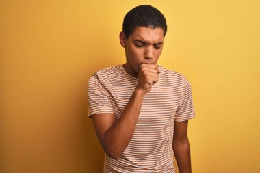 Genç yakışıklı Arap adam izole sarı arka plan üzerinde ayakta çizgili t-shirt giyen rahatsız duygu ve soğuk veya bronşit için bir belirti olarak öksürük. Sağlık kavramı.
