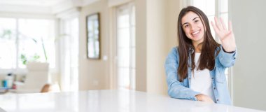 Evde beyaz masada oturan ve kendinden emin ve mutlu gülümserken parmaklarıyla işaret eden genç kadının geniş açılı resmi.