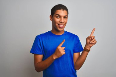 Genç yakışıklı arap adam izole beyaz arka plan üzerinde duran mavi t-shirt giyen gülümseyerek ve yan iki el ve parmakları ile işaret kamera bakıyor.