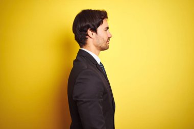 Takım elbise giyen ve kravat giyen genç yakışıklı işadamı yan bakarak izole sarı arka plan üzerinde duran, doğal yüz ve kendine güvenen gülümseme ile profil poz rahatla.