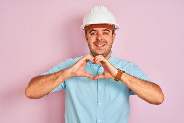 İzole edilmiş pembe arka plan üzerinde duran güvenlik kaskı takan genç mimar, kalp sembolü ve şeklini elleriyle göstererek gülümsüyor. Romantik kavram.