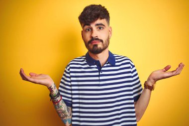 Çizgili polo giyen dövmegiyen genç adam izole sarı arka plan üzerinde duran habersiz ve kolları ve elleri ile karışık ifade kaldırdı. Şüphe kavramı.