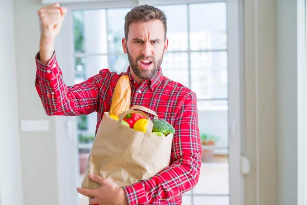 Handsome man holding groceries bag annoyed and frustrated shouting with anger, crazy and yelling with raised hand, anger concept