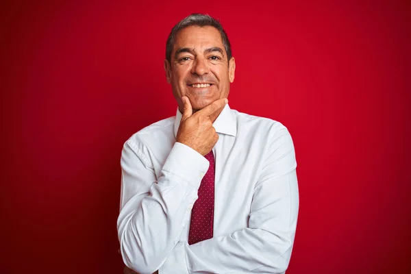 Handsome middle age businessman standing over isolated red background looking confident at the camera smiling with crossed arms and hand raised on chin. Thinking positive.