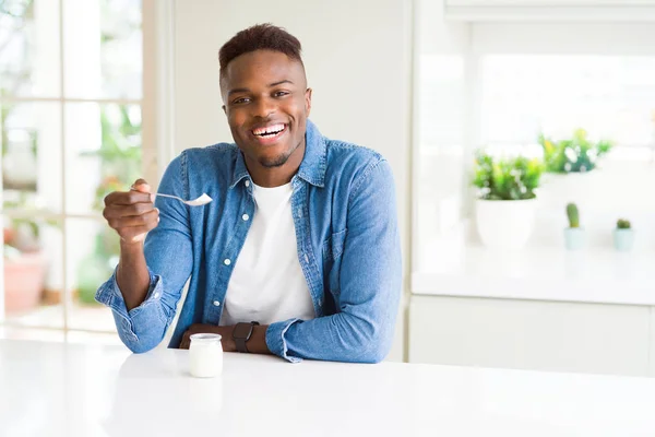 African american man eating healthy natural yogurt with a spoon with a happy face standing and smiling with a confident smile showing teeth