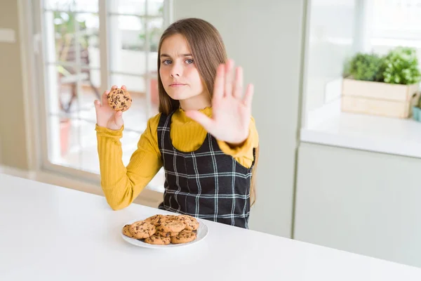 Beautiful young girl kid eating chocolate chips cookies with open hand doing stop sign with serious and confident expression, defense gesture