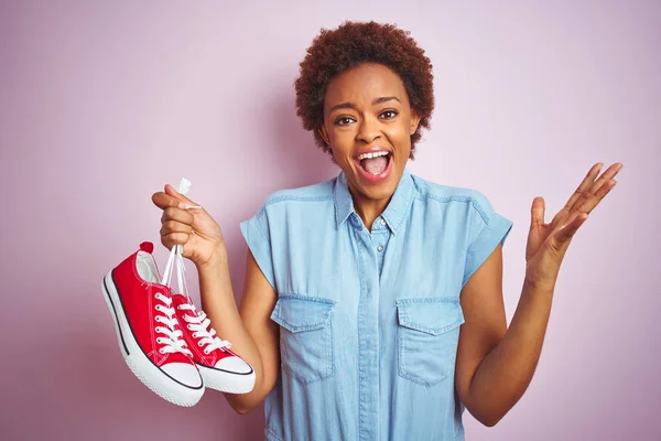 African american woman holding red casual sneakers over pink isolated background very happy and excited, winner expression celebrating victory screaming with big smile and raised hands