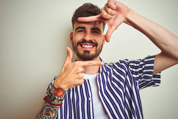 Young man with tattoo wearing striped shirt standing over isolated white background smiling making frame with hands and fingers with happy face. Creativity and photography concept.