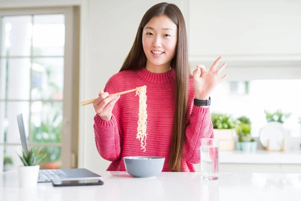 Güzel Asyalı kadın dizüstü bilgisayar kullanarak çalışan ve parmaklarıyla ok işareti yapıyor Asya noodle yeme, mükemmel sembolü