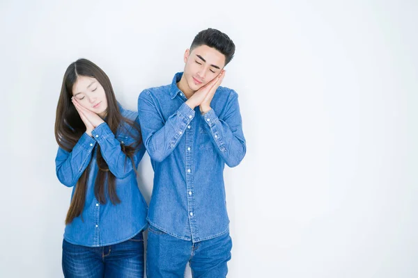 Beautiful young asian couple over white isolated background sleeping tired dreaming and posing with hands together while smiling with closed eyes.