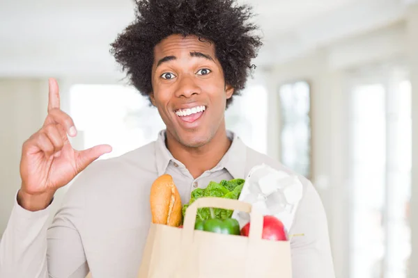 African American man holding groceries bag with fresh vegetables at home surprised with an idea or question pointing finger with happy face, number one