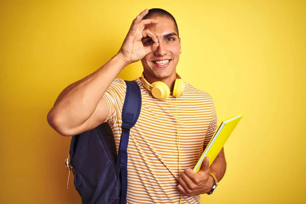 黄色の孤立した背景の上にヘッドフォンとバックパックを身に着けている若い学生の男は 指を通して目に手でOkサインをやって笑顔で幸せな顔をしています — ストック写真