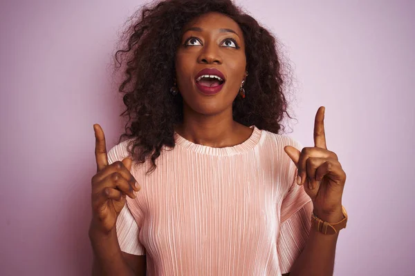İzole pembe arka plan üzerinde duran t-shirt giyen genç Afro-Amerikan kadın şaşırttı ve yukarı bakarak ve parmakları ile işaret ve kolları kaldırdı şaşırttı.