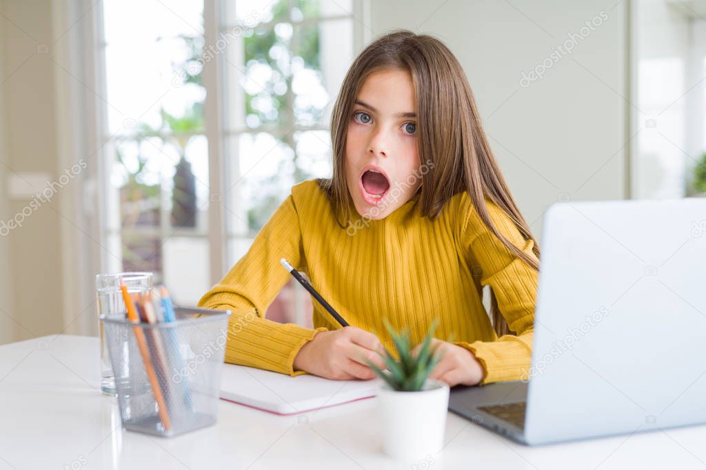 Beautiful young girl studying using computer laptop and writing on notebook scared in shock with a surprise face, afraid and excited with fear expression