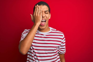 Genç yakışıklı Arap adam izole kırmızı arka plan üzerinde çizgili t-shirt giyen el, yüzünde kendine güvenen gülümseme ve sürpriz duygu ile bir göz kapsayan.