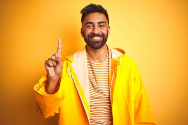 Genç hintli adam izole sarı arka plan üzerinde duran yağmurluk giyen gösteren ve kendinden emin ve mutlu gülümserken parmak bir numara ile işaret.
