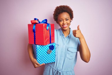 Pembe izole arka plan üzerinde doğum günü hediyeleri tutan Afrikalı Amerikalı kadın ok işareti yapıyor büyük bir gülümseme ile mutlu, parmakları ile başparmak, mükemmel işareti