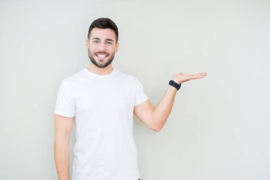 Neşeli sunulması ve kameraya bakarak avucunun ile işaret gülümseyen izole arka plan üzerinde rahat beyaz t-shirt giyen genç yakışıklı adam.