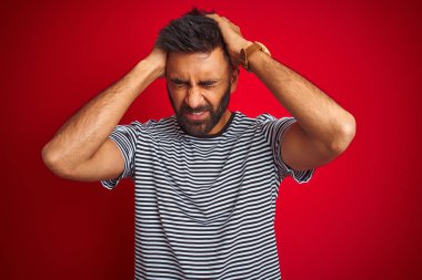 Genç yakışıklı hintli adam izole kırmızı arka plan üzerinde lacivert çizgili t-shirt giyen baş ağrısı umutsuz muzdarip ve stresli çünkü ağrı ve migren. Eller baş üstünde.