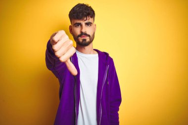 Izole sarı arka plan üzerinde spor mor sweatshirt giyen dövme giyen genç adam mutsuz ve kızgın gösteren reddi ve başparmak aşağı jest ile olumsuz görünümlü. Kötü ifade.