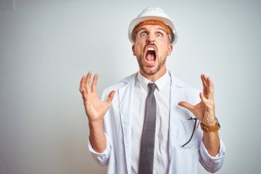 Genç yakışıklı mühendis adam izole arka plan deli ve deli bağırarak üzerinde güvenlik kask giyen ve agresif ifade ve kollar kaldırdı ile bağırıyor. Hayal kırıklığı kavramı.