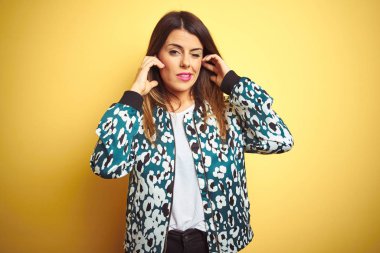 Genç güzel kadın yüksek sesle müzik sesi için rahatsız ifade ile parmakları ile kulakları kapsayan sarı izole arka plan üzerinde rahat ceket giyiyor. Sağır kavramı.