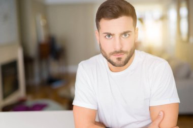 Ev şüpheci ve sinir, çapraz kolları ile yüz üzerinde onaylamaz ifade rahat beyaz t-shirt giyen genç yakışıklı adam. Negatif kişi.