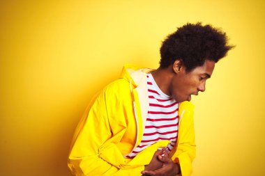 Afro saçlı Afro saçlı afro-amerikan adam mide üzerinde el ile izole sarı arka plan üzerinde duran yağmurluk giyen çünkü hazımsızlık, ağrılı hastalık duygusu. Ağrı kavramı.