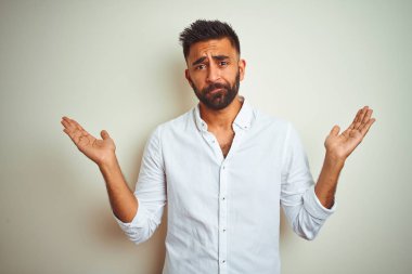 Genç hintli adam zarif gömlek giyen izole beyaz arka plan üzerinde duran habersiz ve kolları ve elleri kaldırdı ile karışık ifade. Şüphe kavramı.