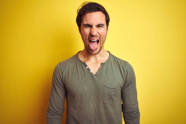 Genç yakışıklı bir adam günlük tişört giyip izole edilmiş sarı arka planda dikiliyor ve komik bir ifadeyle dilini dışarı çıkarıyor. Duygu kavramı.