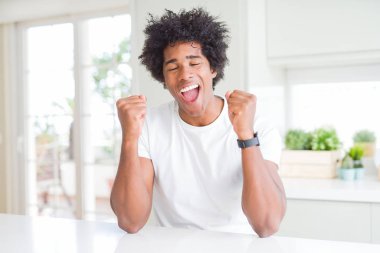 Evde oturan rahat beyaz tişört giyen genç Afro-Amerikan adam başarı için heyecanlı kolları kaldırdı ve gözleri kapalı zafer gülümseyerek kutluyor. Kazanan kavramı.