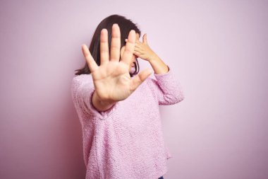 Genç güzel esmer kadın pembe izole arka plan üzerinde bir kazak giyen elleri ile gözleri kaplayan ve üzgün ve korku ifadesi ile stop jest yapıyor. Utanmış ve olumsuz kavram.