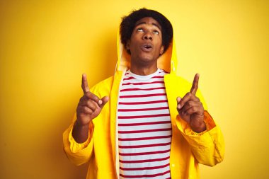 Afro saçlı afro saçlı, izole edilmiş sarı arka plan üzerinde duran davlumbaz yağmurluk giyen Afrikalı adam şaşırmış ve şaşırmış yukarı bakarak ve parmakları ile işaret ve kolları kaldırdı.