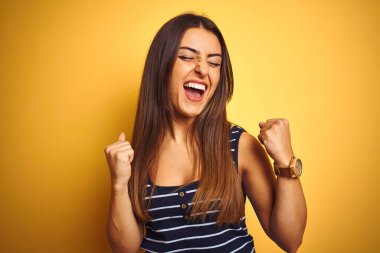 İzole sarı arka plan üzerinde duran çizgili t-shirt giyen genç güzel kadın çok mutlu ve heyecanlı kolları kaldırdı ile kazanan jest yapıyor, gülümseyerek ve başarı için çığlık. Kutlama konsepti.