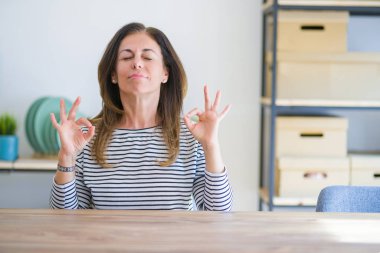 Orta yaş yaşlı kadın evde masada oturan rahat ve gözleri parmakları ile meditasyon jest yapıyor kapalı gülümseyerek. Yoga konsepti.
