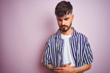 Dövmesi olan genç adam çizgili gömlek giyen mide üzerinde el ile izole pembe arka plan üzerinde duran çünkü mide, ağrılı hastalık kötü hissediyorum. Ağrı kavramı.