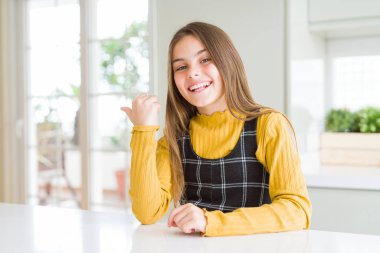 Genç güzel sarışın çocuk kız evde rahat sarı kazak giyen mutlu yüz görünümlü gülümseyerek ve başparmak ile yan işaret.