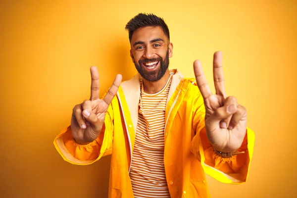 Young indian man wearing raincoat standing over isolated yellow background smiling looking to the camera showing fingers doing victory sign. Number two.