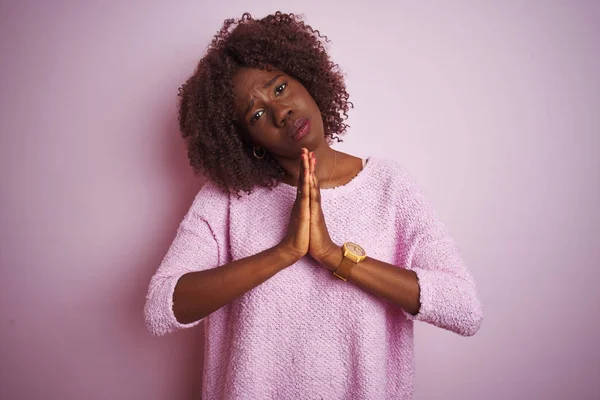 Young african afro woman wearing sweater standing over isolated pink background begging and praying with hands together with hope expression on face very emotional and worried. Asking for forgiveness. Religion concept.