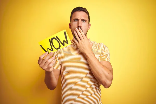 Young man holding wow text banner over yellow isolated background cover mouth with hand shocked with shame for mistake, expression of fear, scared in silence, secret concept