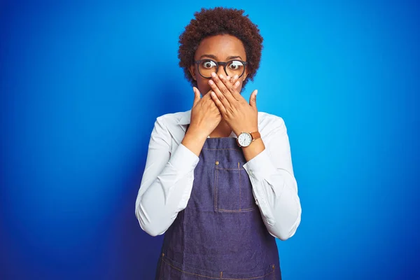 Young african american woman shop owner wearing business apron over blue background shocked covering mouth with hands for mistake. Secret concept.