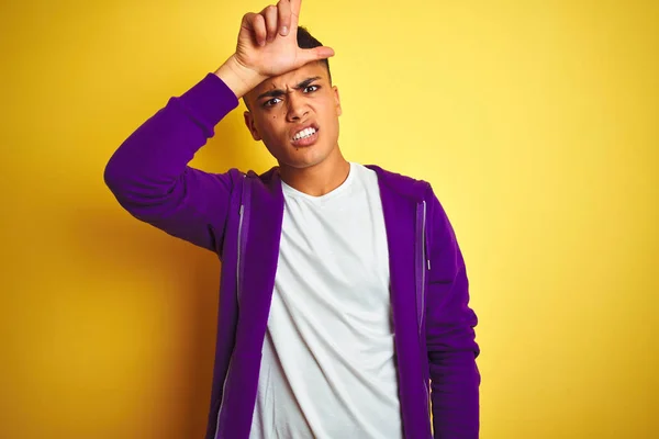 年轻的巴西男子穿着紫色运动衫站在孤立的黄色背景上取笑人用手指在额头做失败者手势嘲笑和侮辱 — 图库照片