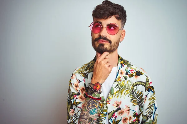 Tatilde dövmeli adam yazlık gömlek güneş gözlüğü takan izole beyaz arka plan üzerinde çene üzerinde el soru, dalgın ifade düşünme. Düşünceli bir yüzle gülümsüyor. Şüphe kavramı.