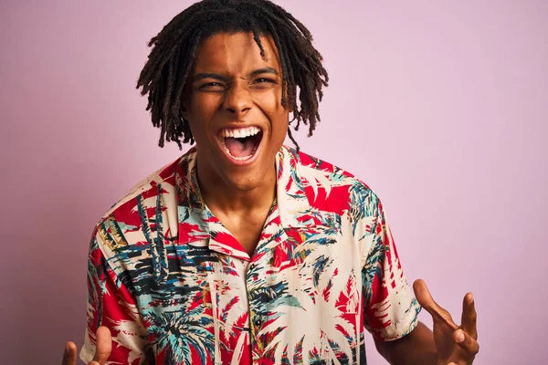 Tatilde dreadlocks ile Afro adam izole pembe arka plan deli ve deli bağırarak ve agresif ifade ve kollar kaldırdı ile bağırıyor üzerinde çiçek gömlek giyiyor. Hayal kırıklığı kavramı.