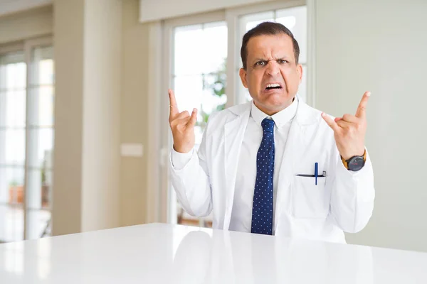 Middle age doctor man wearing medical coat at the clinic shouting with crazy expression doing rock symbol with hands up. Music star. Heavy concept.