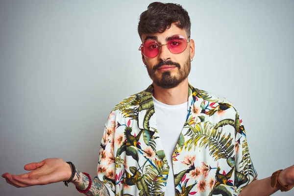 Tatilde dövme ile Adam izole beyaz arka plan üzerinde yazlık gömlek güneş gözlüğü giyen habersiz ve kollar ve eller kaldırdı ile karışık ifade. Şüphe kavramı.