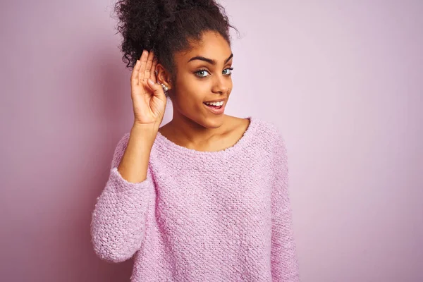 Young african american woman wearing winter sweater standing over isolated pink background smiling with hand over ear listening an hearing to rumor or gossip. Deafness concept.