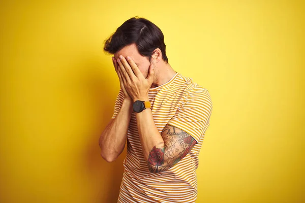 孤立した黄色の背景にストライプのTシャツを着た若いハンサムな男は 泣きながら手で顔を覆う悲しい表情をしています うつ病の概念 — ストック写真